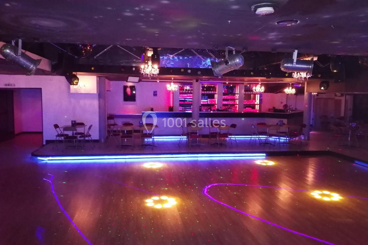 Salle de danse avec piste éclairée par des lumières colorées, bar illuminé en arrière-plan et chaises disposées autour.