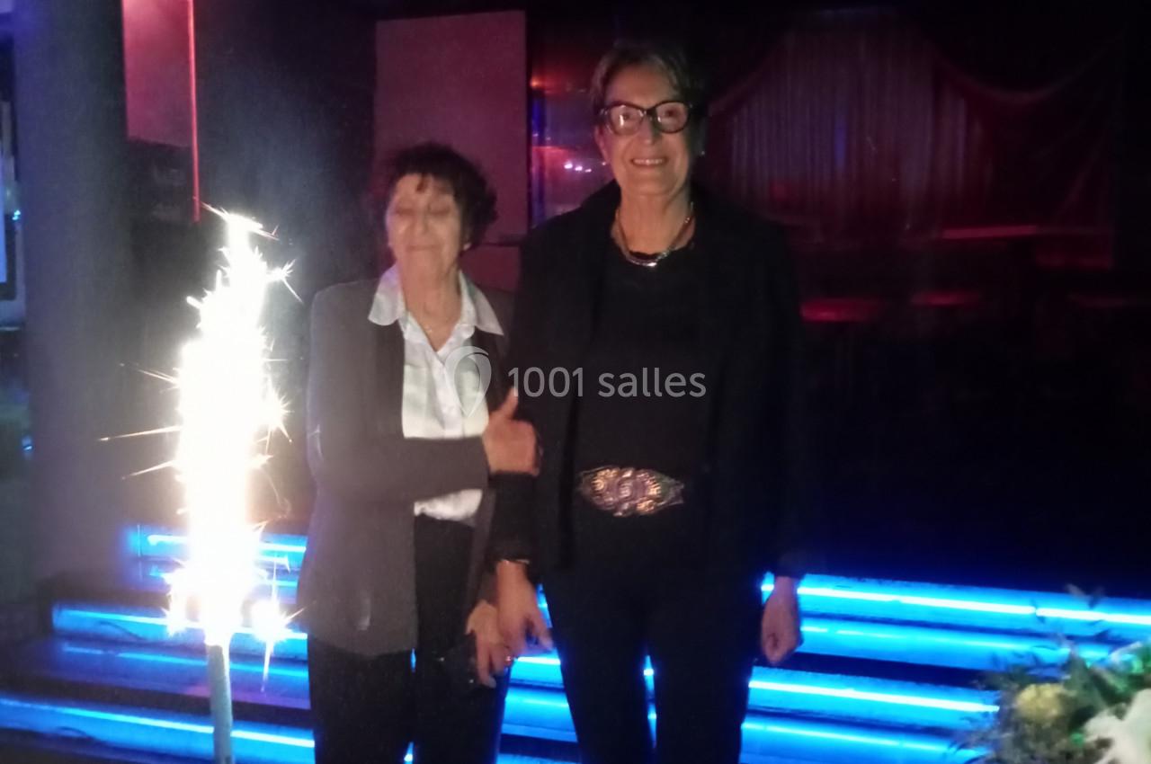 Deux femmes debout dans un environnement sombre près d'un gâteau d'anniversaire avec une bougie scintillante.