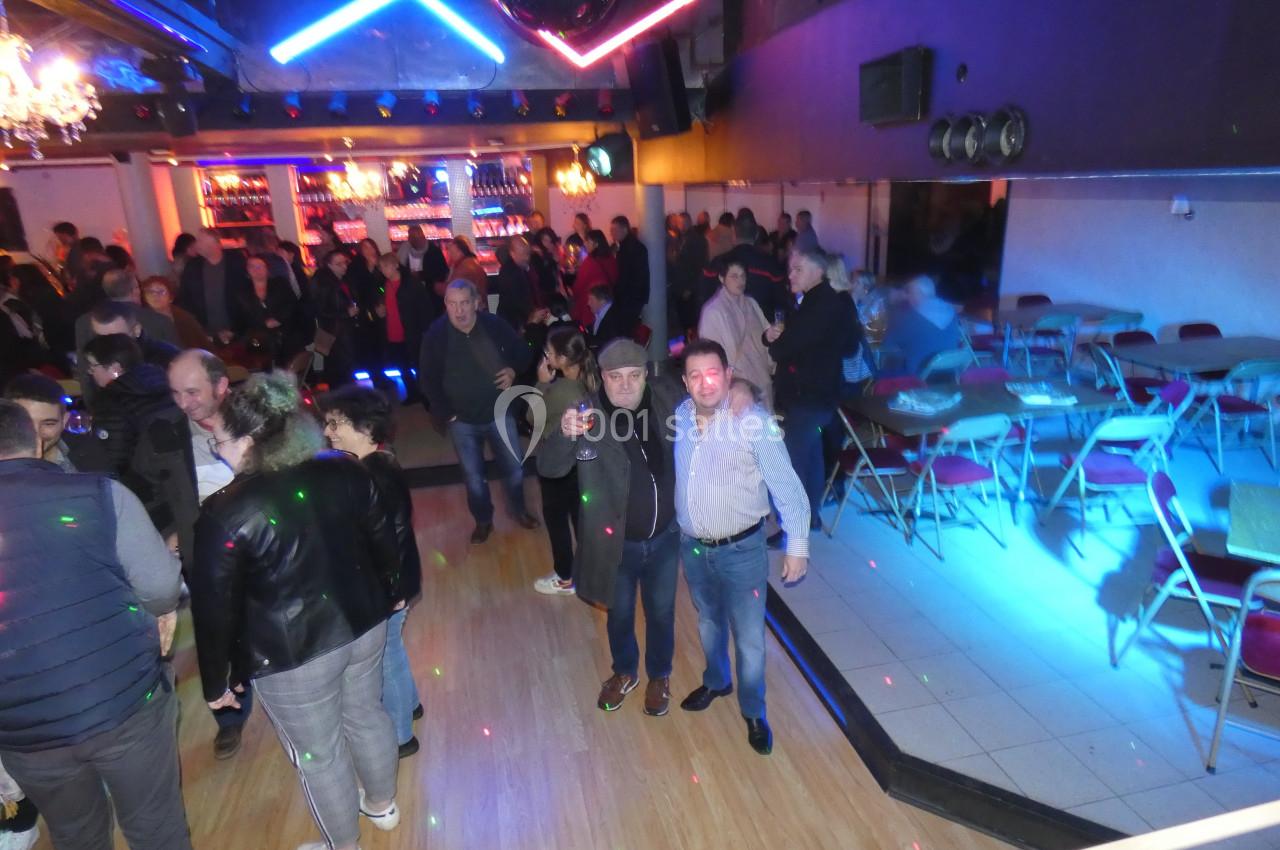 Personnes rassemblées dans une salle de fête éclairée, avec piste de danse et tables sur le côté.
