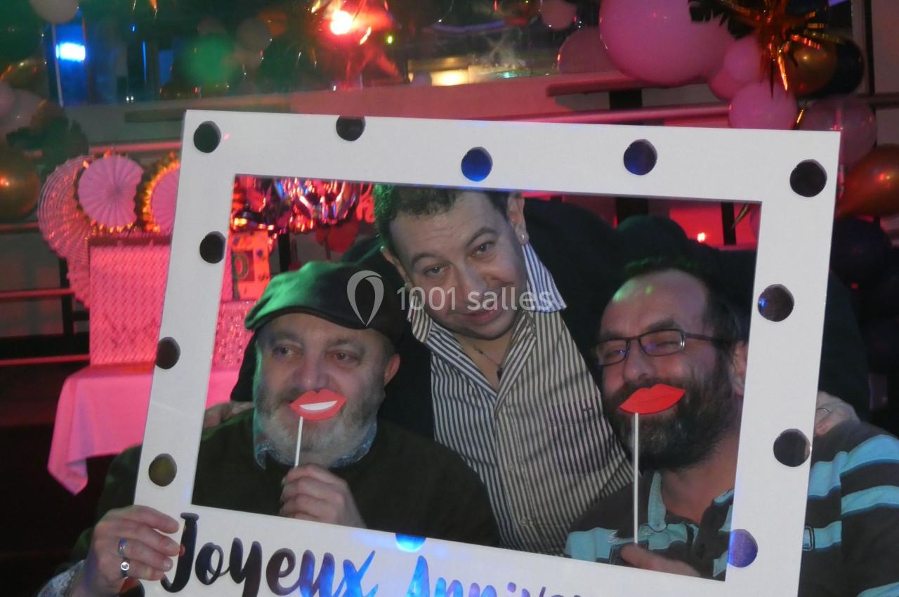 Trois hommes posent avec un cadre ’Joyeux Anniversaire’ et des accessoires amusants dans une ambiance festive.