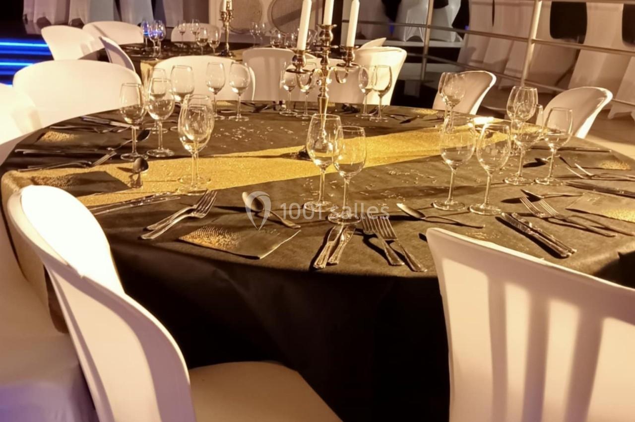 Table ronde élégamment dressée avec nappe noire, chemin de table doré, chandeliers et verres, entourée de chaises blanches.