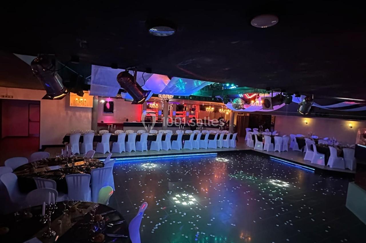 Salle de réception sombre avec piste de danse illuminée, tables dressées et bar éclairé en arrière-plan.