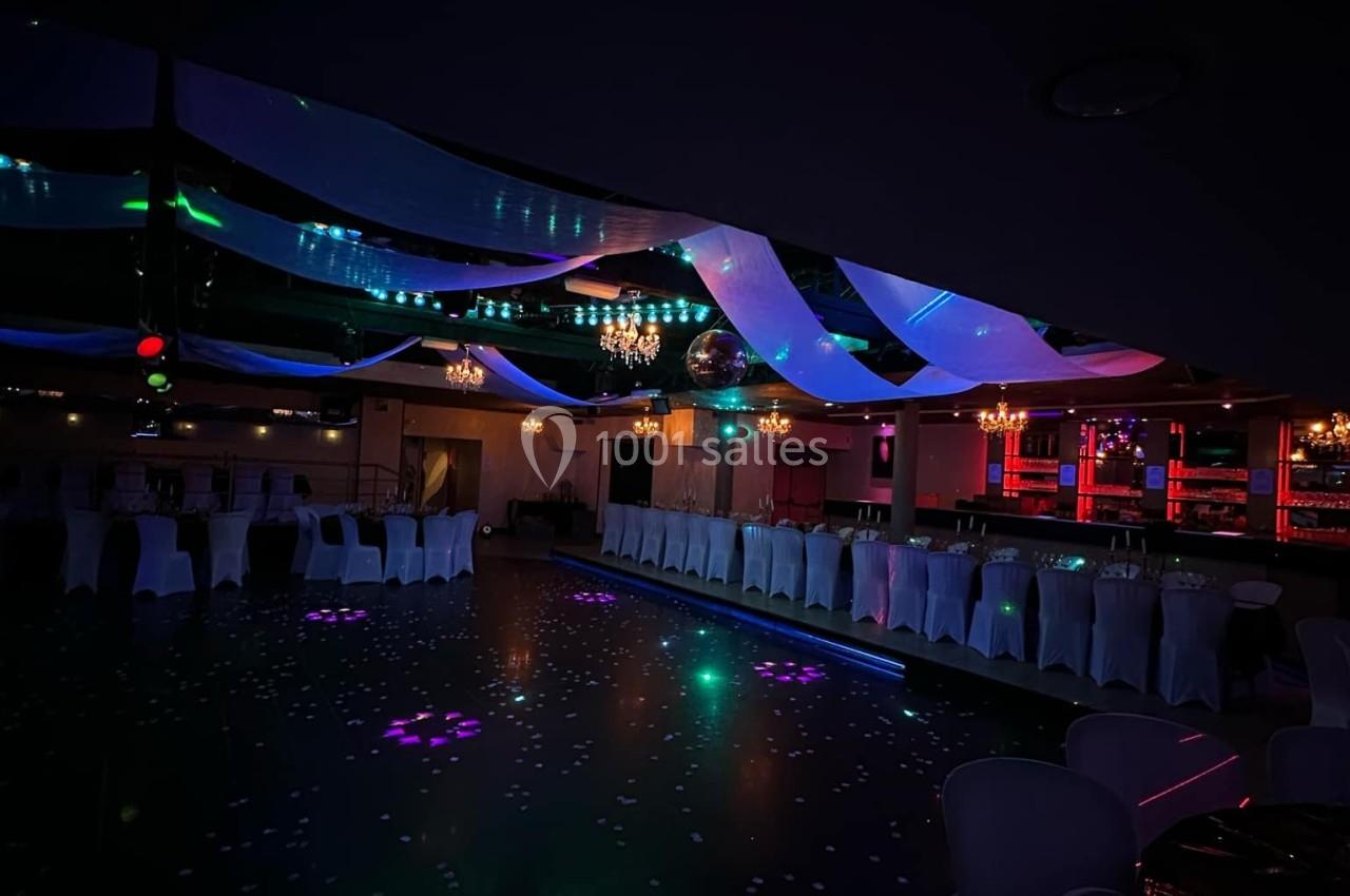 Salle de réception sombre avec éclairage coloré, tables alignées et chaises blanches sous des décorations suspendues.