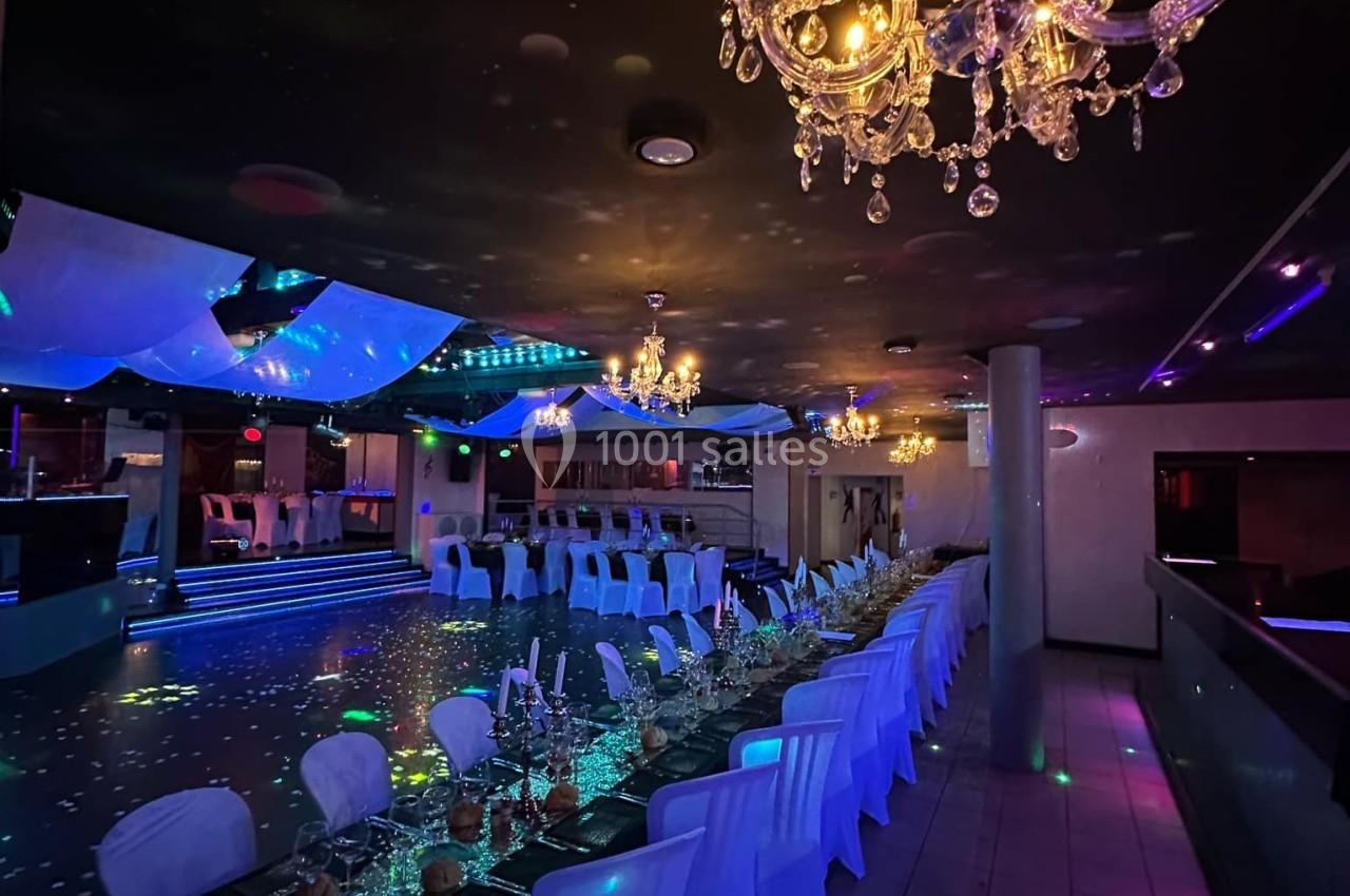 Salle de réception décorée avec des tables alignées, nappes blanches, chandeliers suspendus et éclairage tamisé coloré.