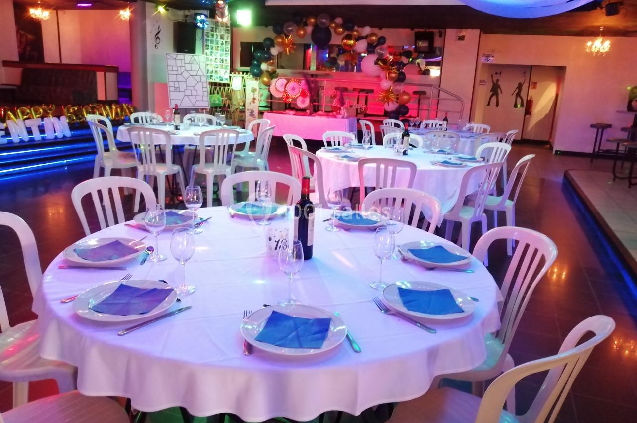 Salle décorée pour un événement, avec des tables rondes dressées, nappes blanches et chaises en plastique.
