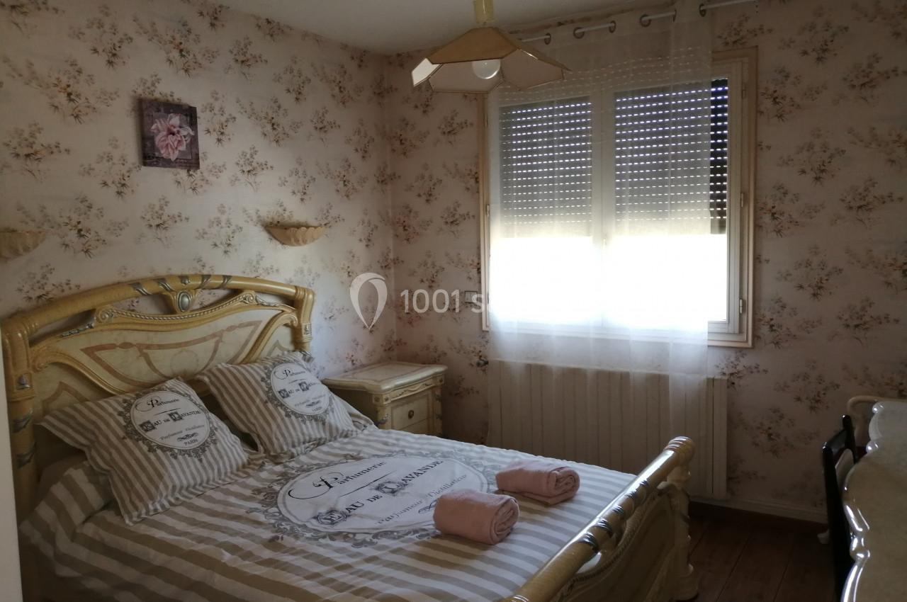 Chambre avec lit double, décor floral, lumière naturelle filtrée par des rideaux et volets partiellement fermés.