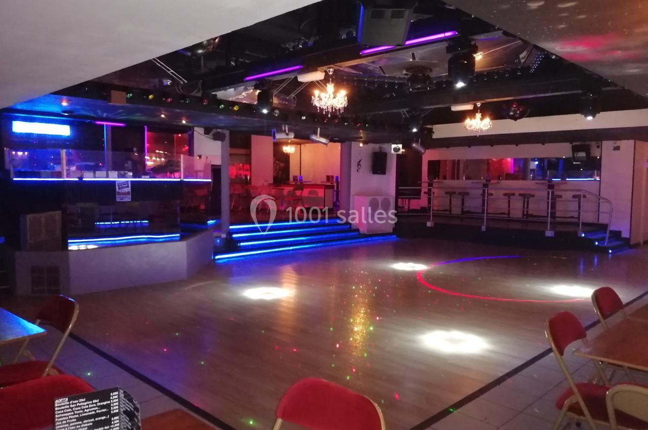 Salle de danse vide avec piste en parquet, éclairages colorés, tables et chaises autour, ambiance de soirée.