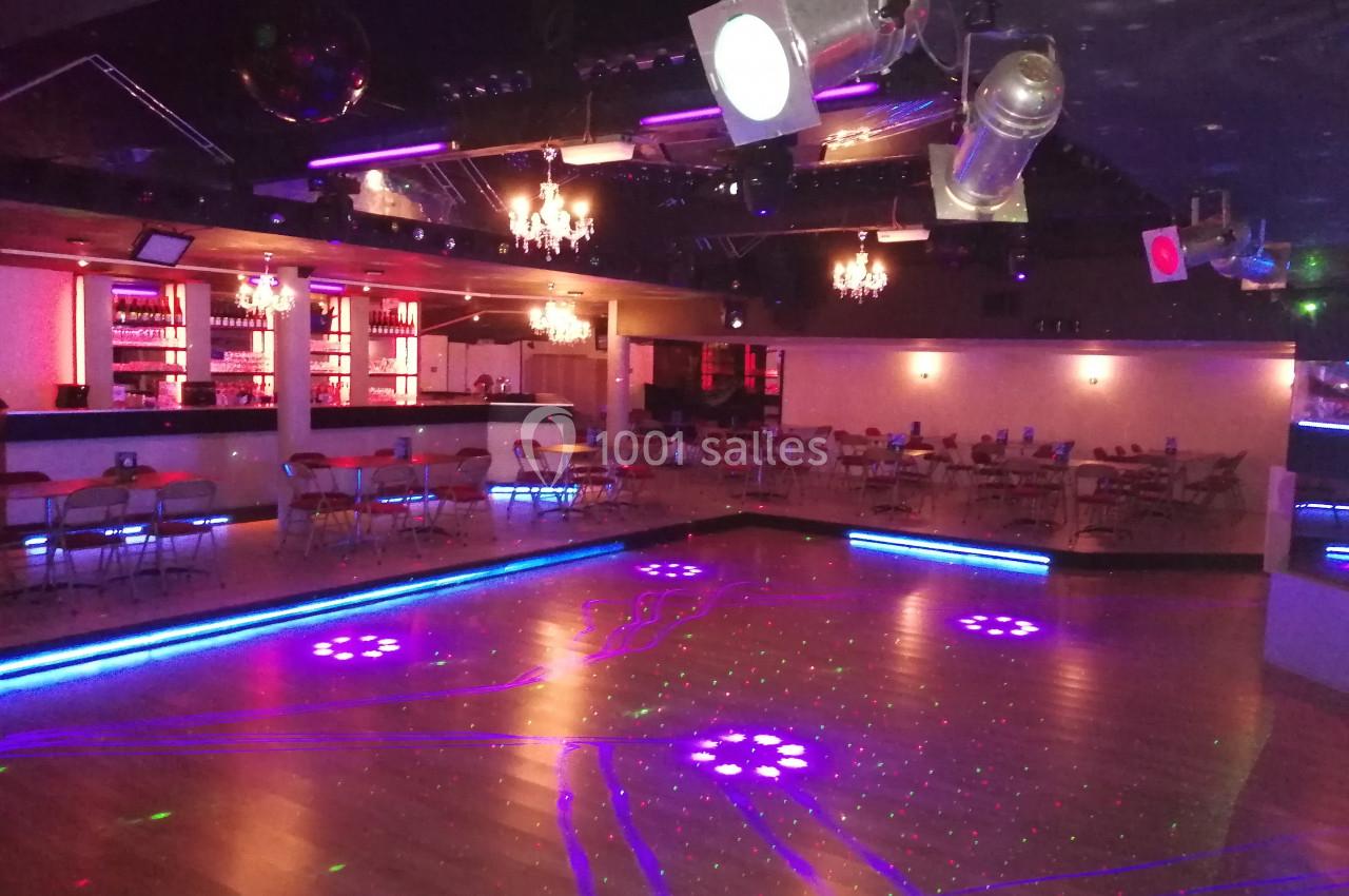 Salle de danse avec éclairage coloré, piste en bois, tables et chaises disposées près d'un bar illuminé.