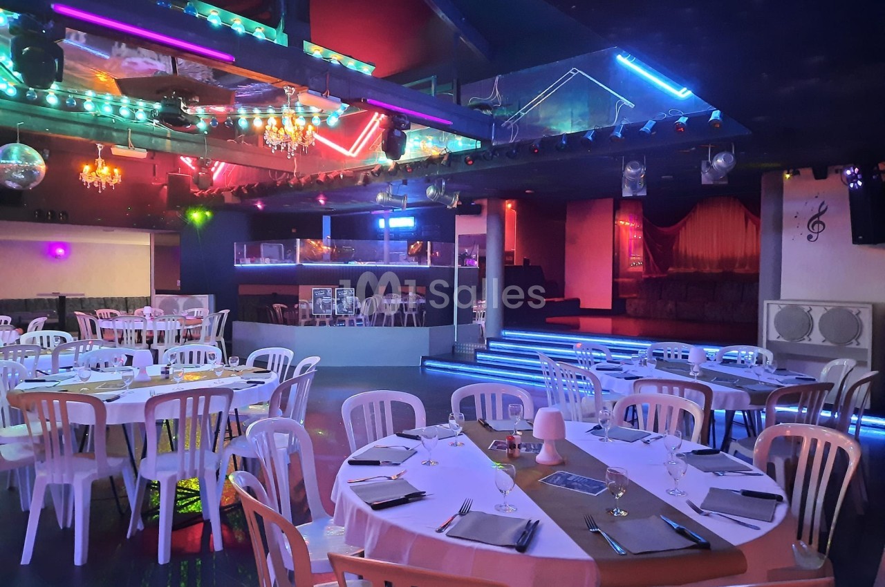 Salle de réception avec tables rondes dressées, chaises blanches et éclairages colorés dans une ambiance festive.