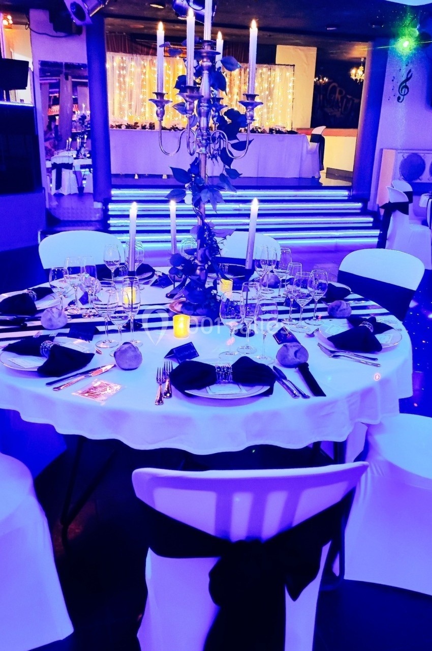 Table élégamment dressée avec nappes blanches, chandeliers, verres et couverts, dans une salle éclairée en lumière bleue.