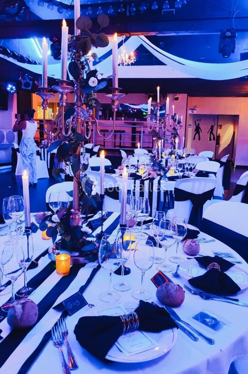 Salle de réception décorée avec des chandeliers, nappes blanches et noires, verres et bougies sur les tables.