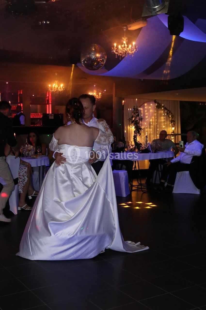 Un couple en tenue de mariage danse dans une salle éclairée par des lumières tamisées et des décorations festives.