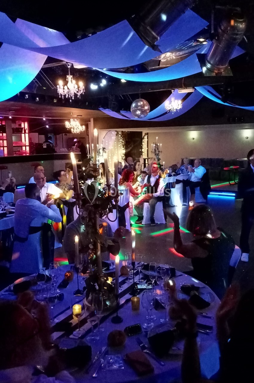 Salle de réception décorée avec des guirlandes et des lumières colorées, des invités assis et dansant.