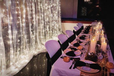 Salle de réception décorée avec des guirlandes et des lumières colorées, des invités assis et dansant.