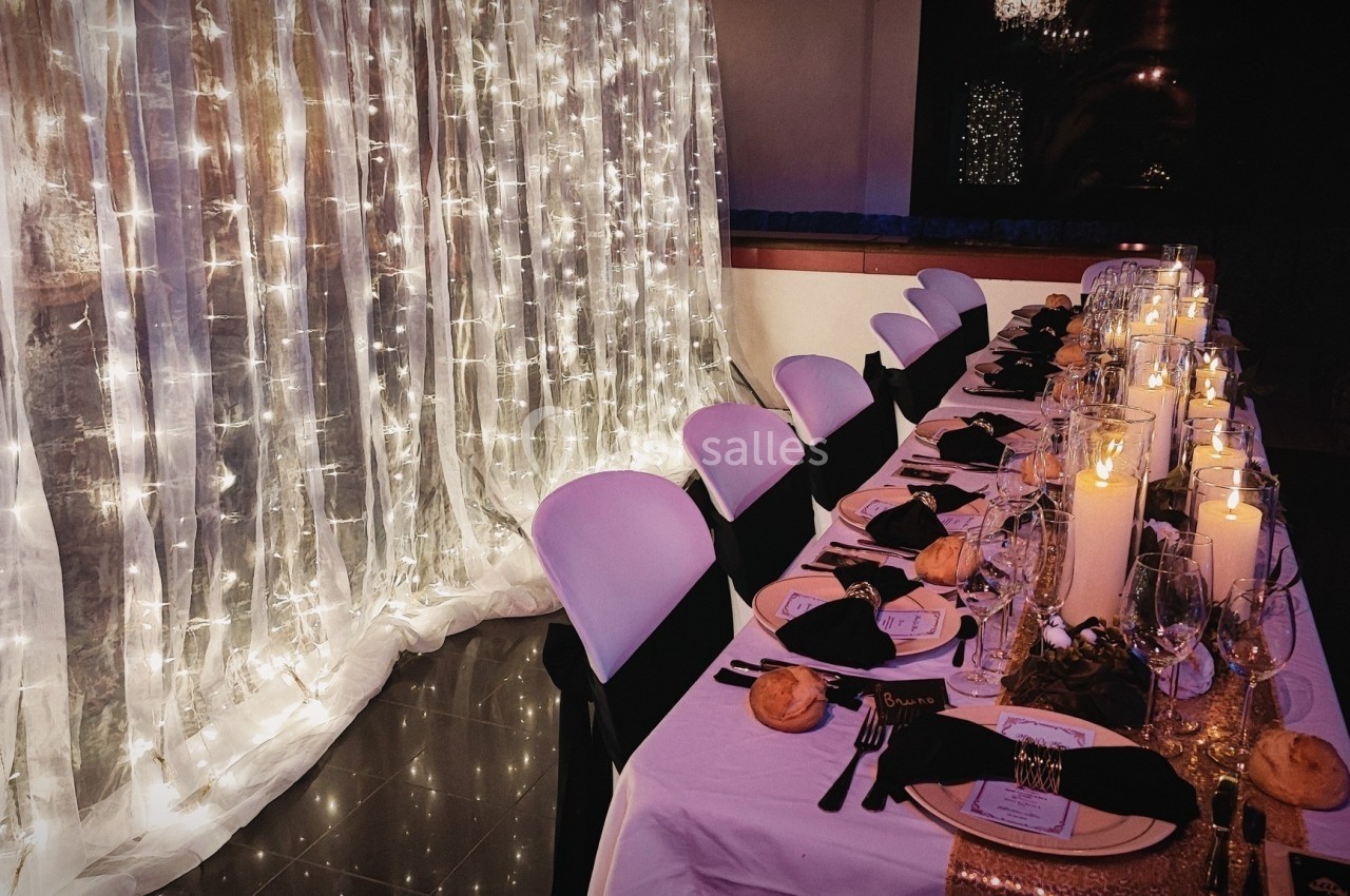 Table élégamment dressée avec nappes blanches, bougies, serviettes noires et guirlandes lumineuses en arrière-plan.