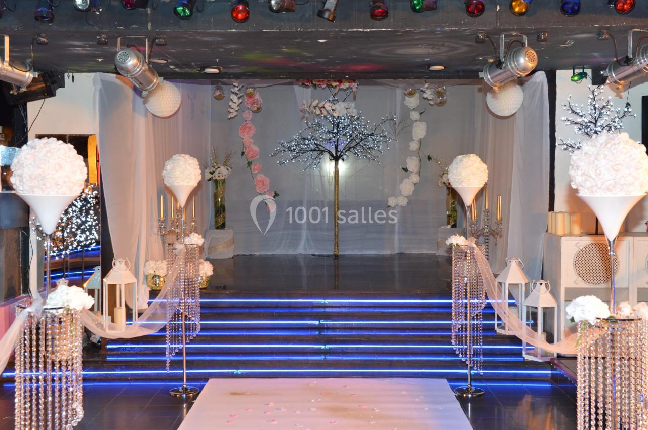 Décoration de salle de réception avec allée centrale, fleurs blanches, guirlandes lumineuses et fond en voile translucide.