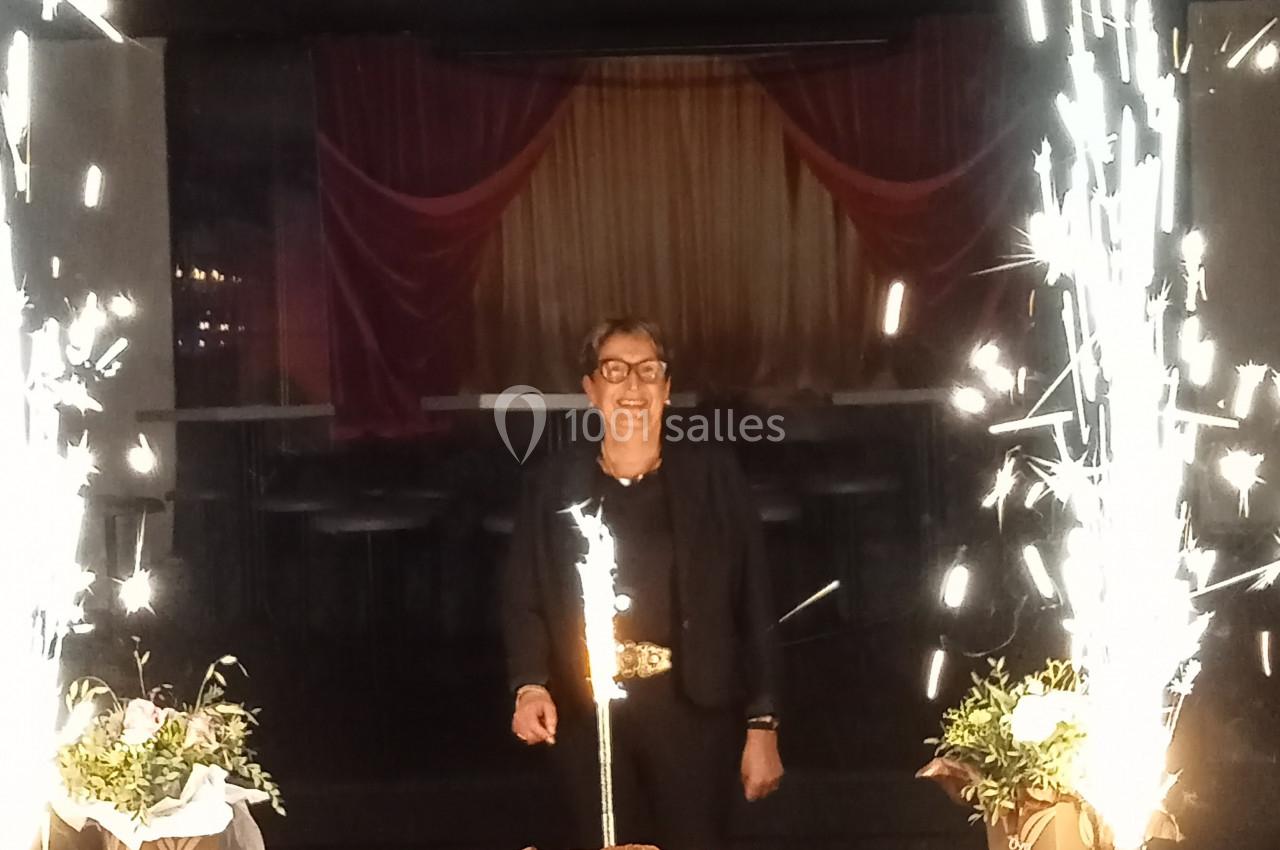 Une personne souriante debout derrière un gâteau d'anniversaire entouré de fontaines lumineuses et de décorations.