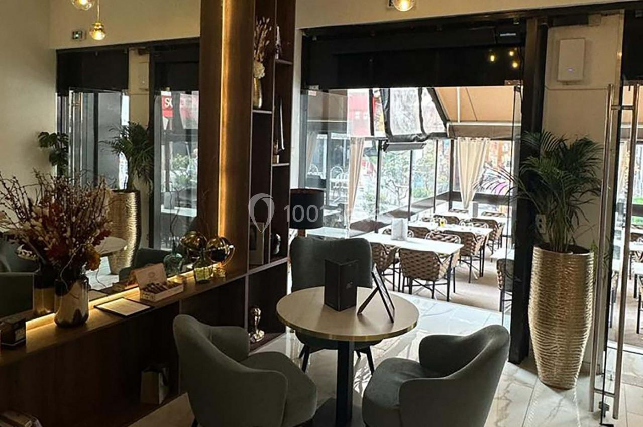 Intérieur d'un café moderne avec tables, chaises rembourrées, plantes décoratives et vue sur une terrasse extérieure.