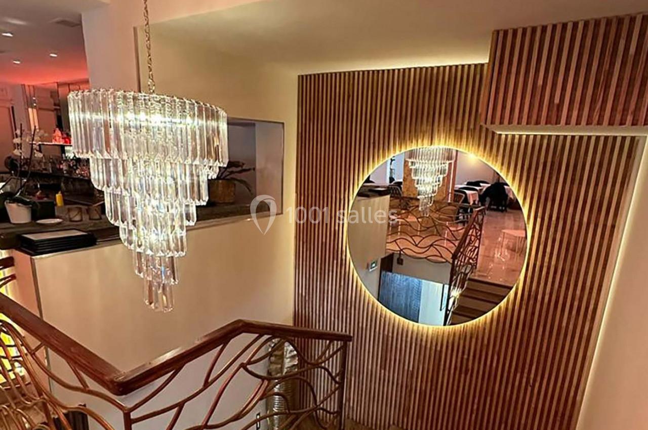 Escalier intérieur avec rampe dorée, miroir rond éclairé et lustre en verre suspendu dans un décor moderne.