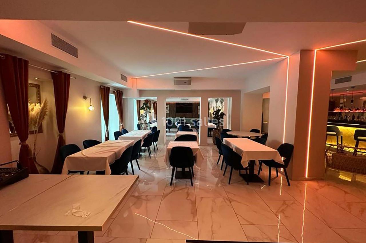 Salle de restaurant moderne avec tables dressées, chaises noires, éclairage tamisé et bar lumineux en arrière-plan.