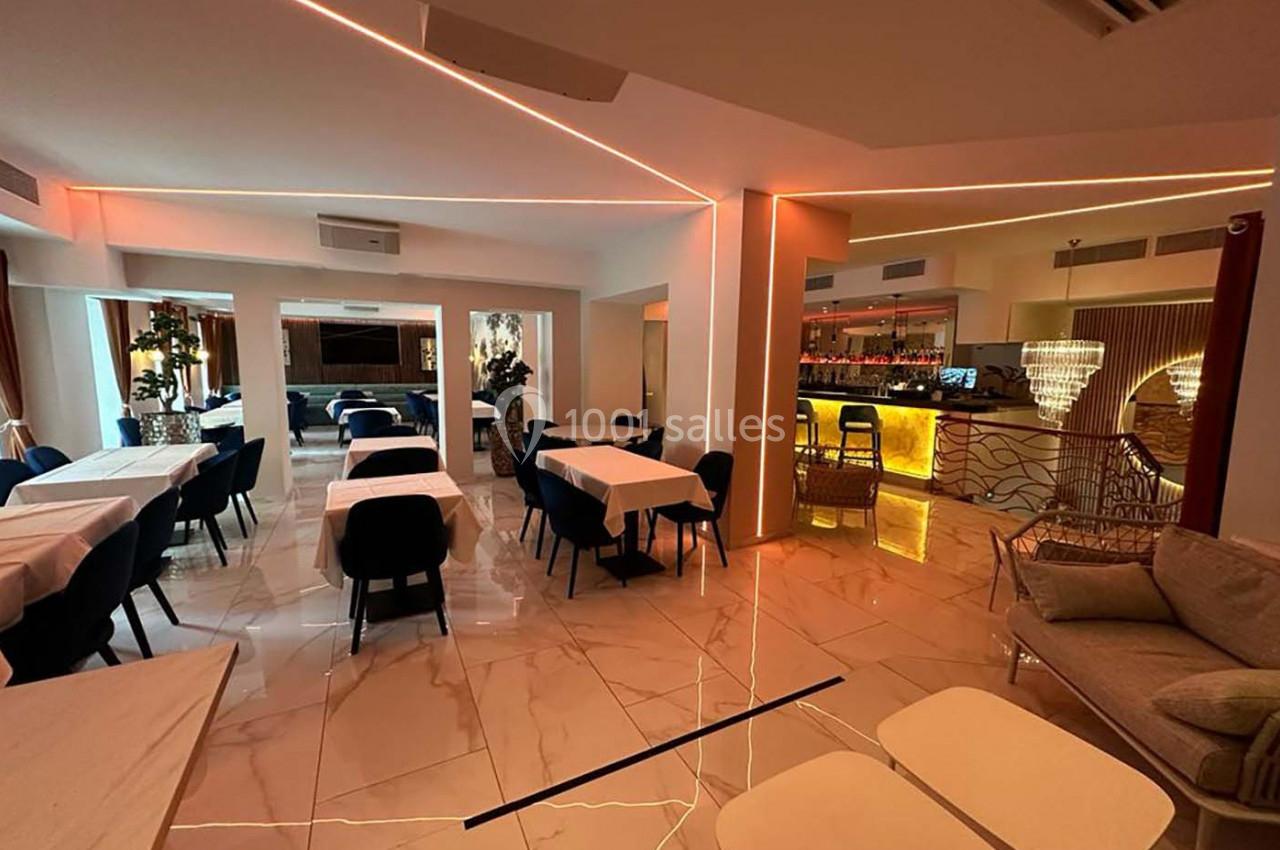 Salle de restaurant moderne avec tables dressées, chaises noires, éclairage chaleureux et bar illuminé en arrière-plan.