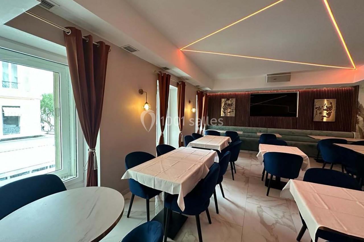 Salle de restaurant moderne avec tables dressées, chaises bleues, rideaux rouges et éclairage géométrique au plafond.