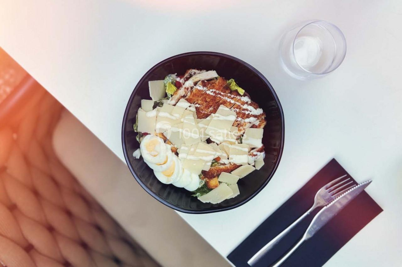 Salade César dans un bol noir avec poulet, œuf, parmesan et sauce, posée sur une table blanche avec couverts.