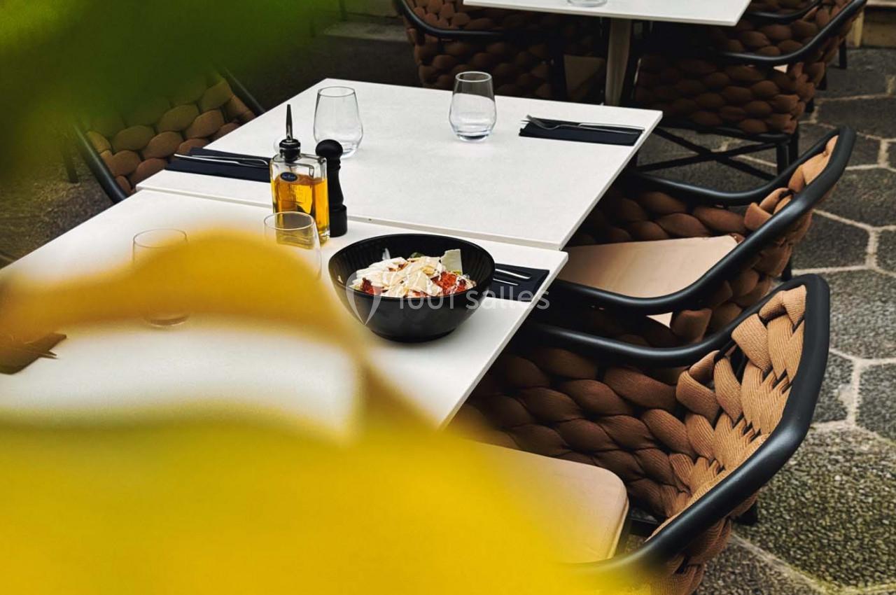 Table de restaurant en extérieur avec des chaises tressées, un plat de pâtes et des condiments sur la table.