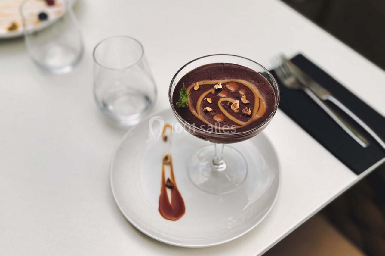 Dessert au chocolat dans une coupe, décoré de noisettes et d'un filet de sauce, servi sur une assiette blanche.