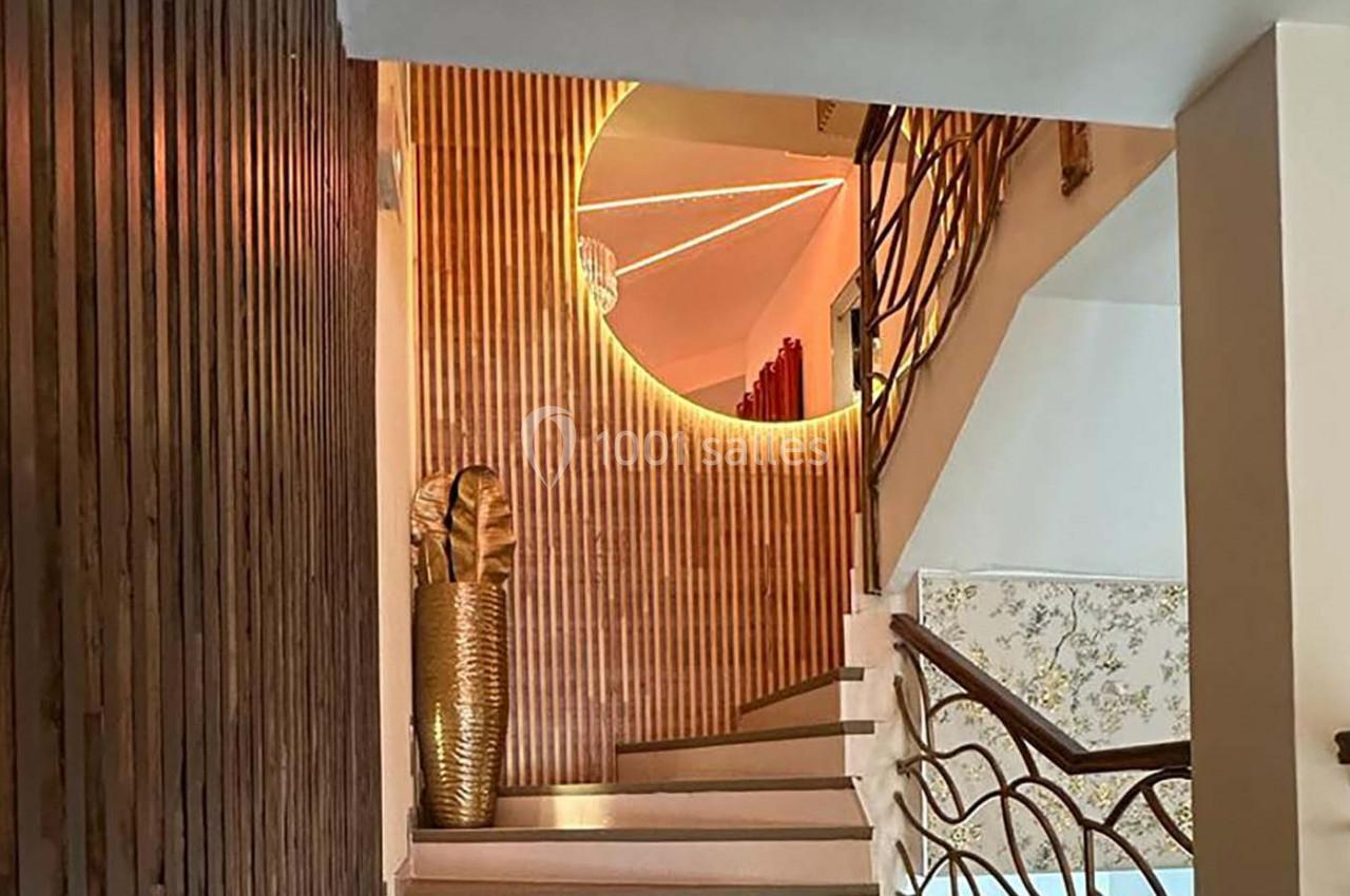 Escalier intérieur avec mur en bois, miroir rond rétroéclairé et vase doré décoratif au pied des marches.