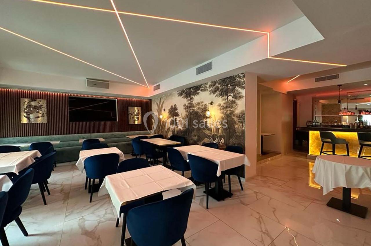 Salle de restaurant moderne avec tables dressées, chaises bleues, éclairage néon et décor mural végétal.