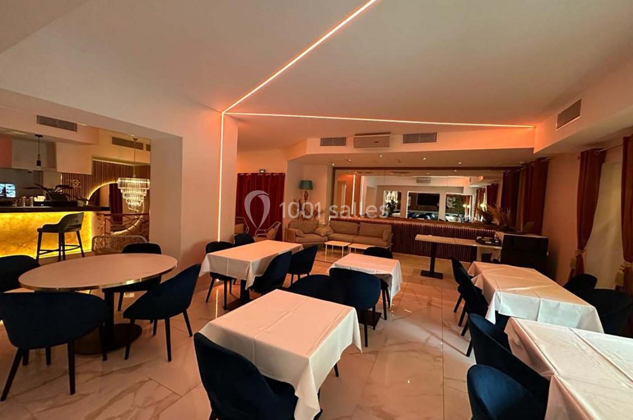 Salle de restaurant moderne avec tables dressées, chaises bleues, éclairage tamisé et bar illuminé en arrière-plan.