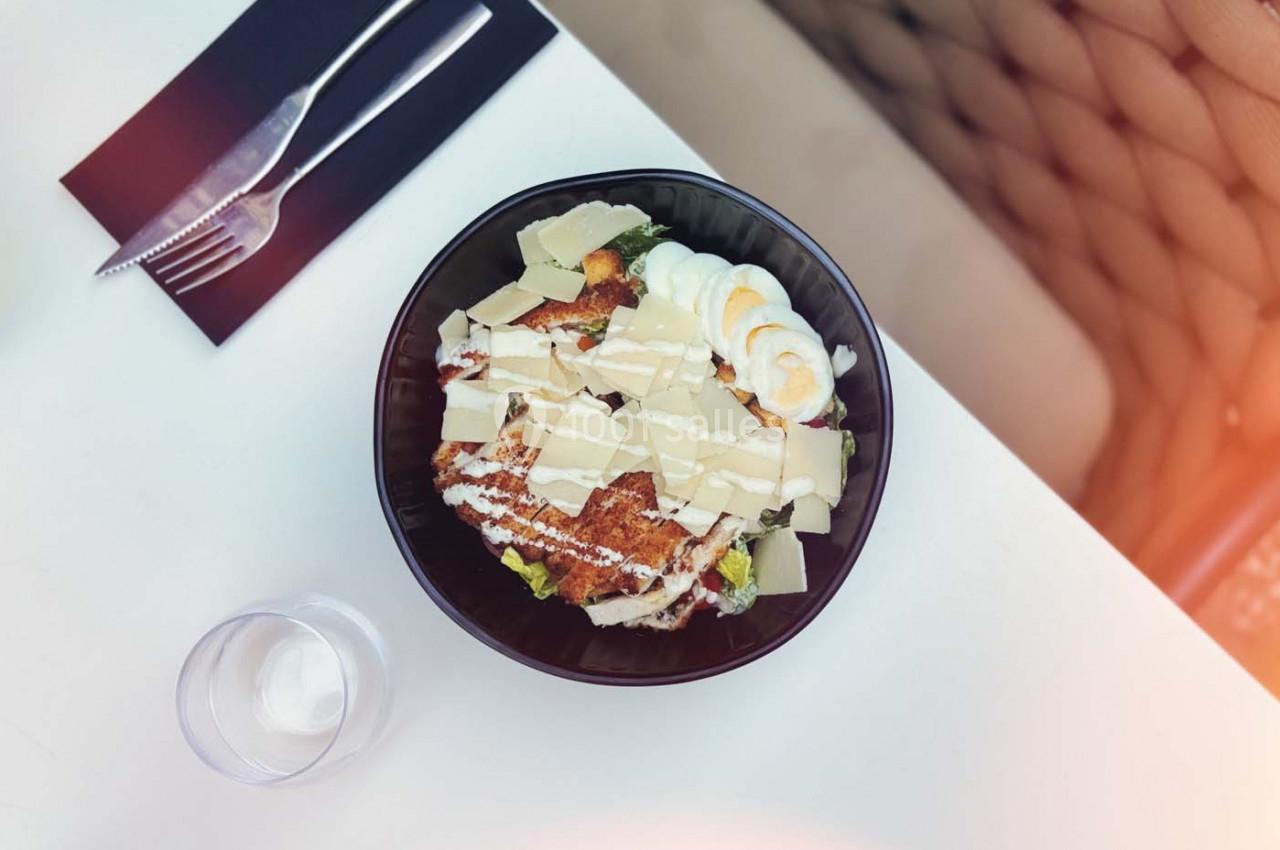 Salade César dans un bol noir avec poulet grillé, œufs durs, copeaux de parmesan et laitue, sur une table blanche.