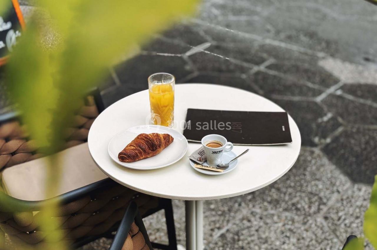 Table ronde en extérieur avec un croissant, un café, un jus d'orange et un menu posé sur une chaise en arrière-plan.
