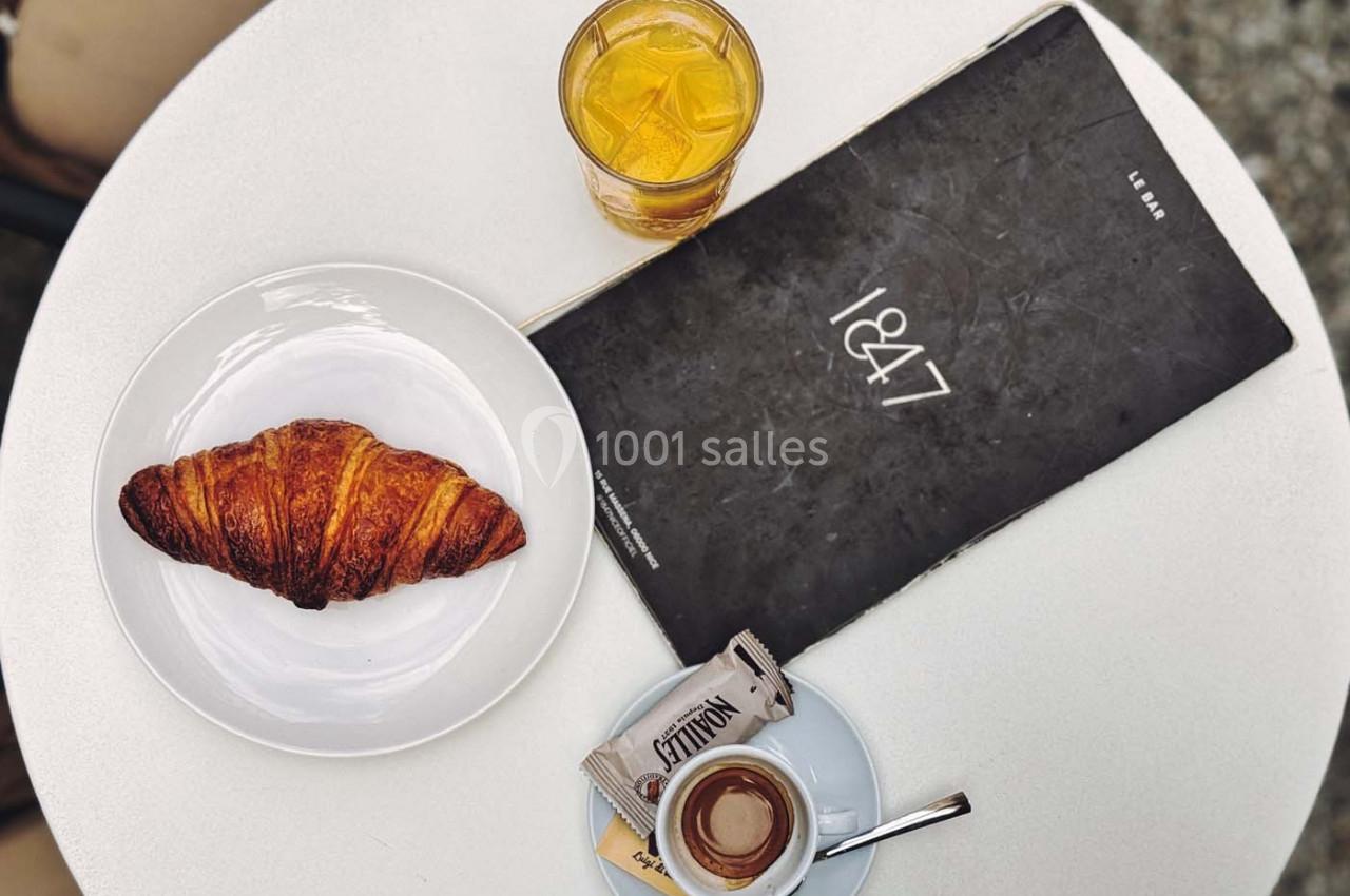 Croissant sur une assiette, café avec un sachet de sucre, jus d'orange et menu sur une table ronde blanche.