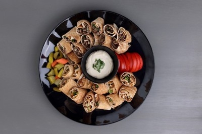 Assiette de wraps garnis autour d'un bol de sauce blanche, accompagnée de légumes marinés et de tranches de tomate.