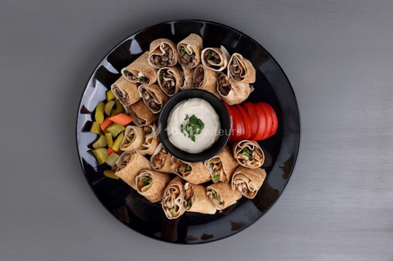 Assiette de wraps garnis autour d'un bol de sauce blanche, accompagnée de légumes marinés et de tranches de tomate.