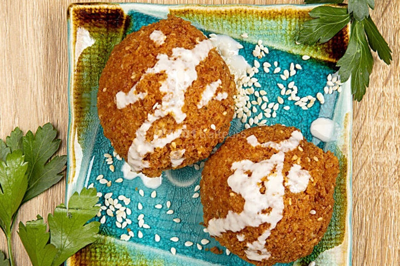 Deux falafels nappés de sauce blanche et parsemés de graines de sésame, présentés sur une assiette colorée avec du persil.