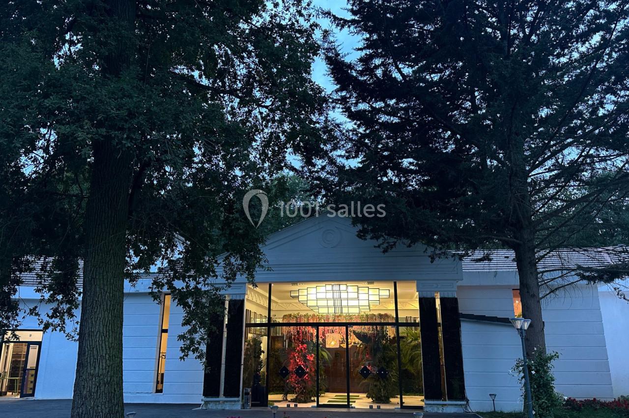 Entrée d'un bâtiment blanc entouré d'arbres, avec des portes vitrées illuminées par une lumière intérieure.