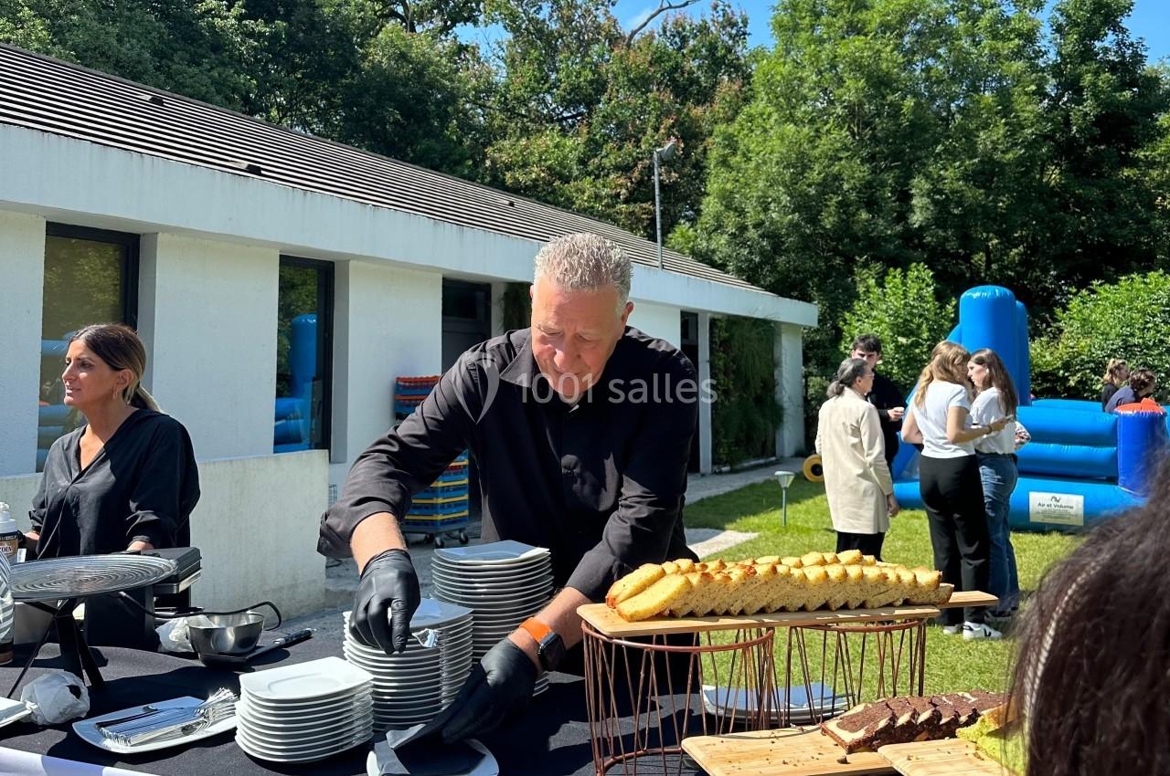 Un homme sert des assiettes sur une table de buffet en extérieur, avec des invités et un château gonflable en arrière-plan.