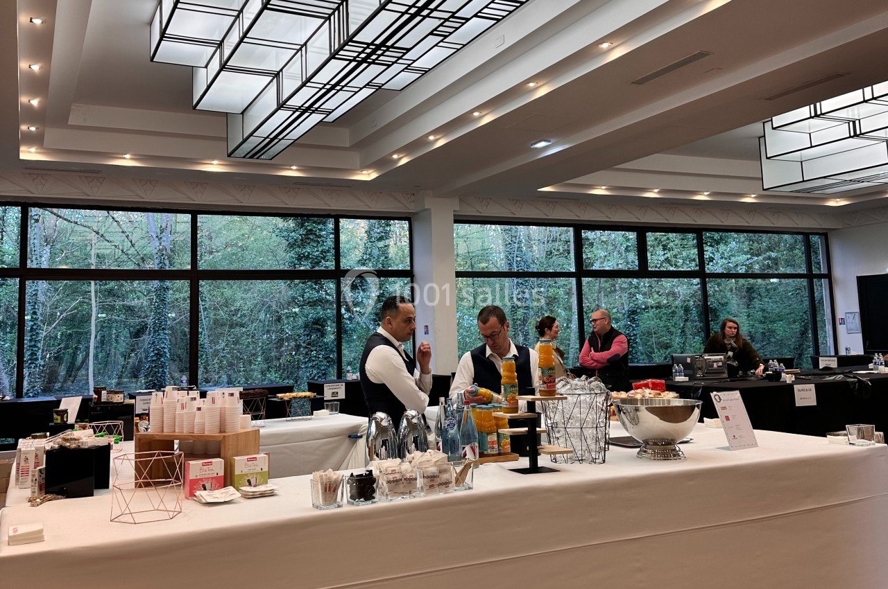 Buffet avec boissons, snacks et serveurs dans une salle lumineuse avec de grandes baies vitrées donnant sur un espace arboré.