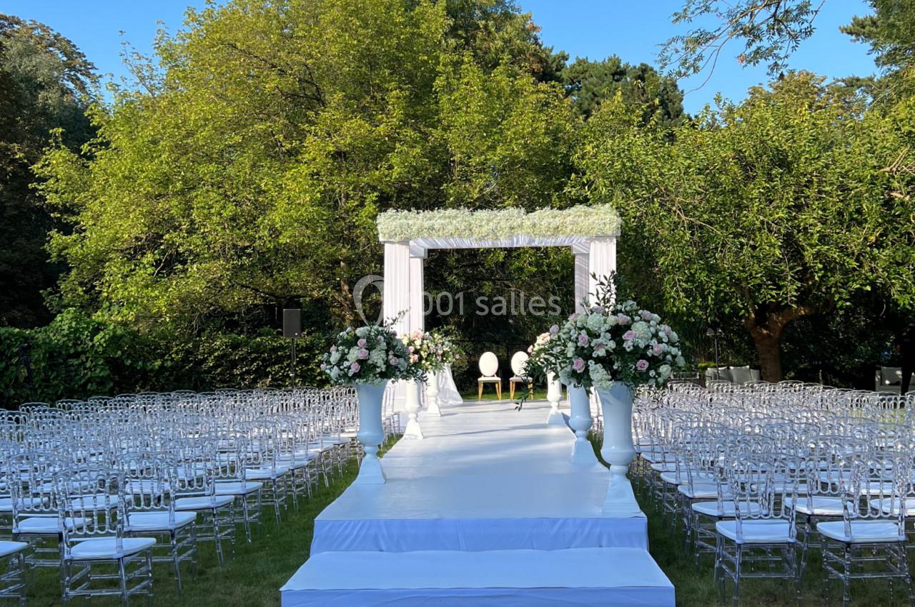 Allée centrale menant à une arche de mariage décorée de fleurs blanches et roses, entourée de chaises transparentes en…