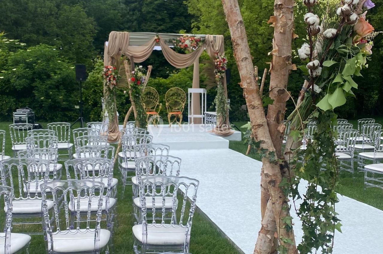 Allée centrale menant à une arche de mariage décorée de fleurs et de tissus, entourée de chaises transparentes.