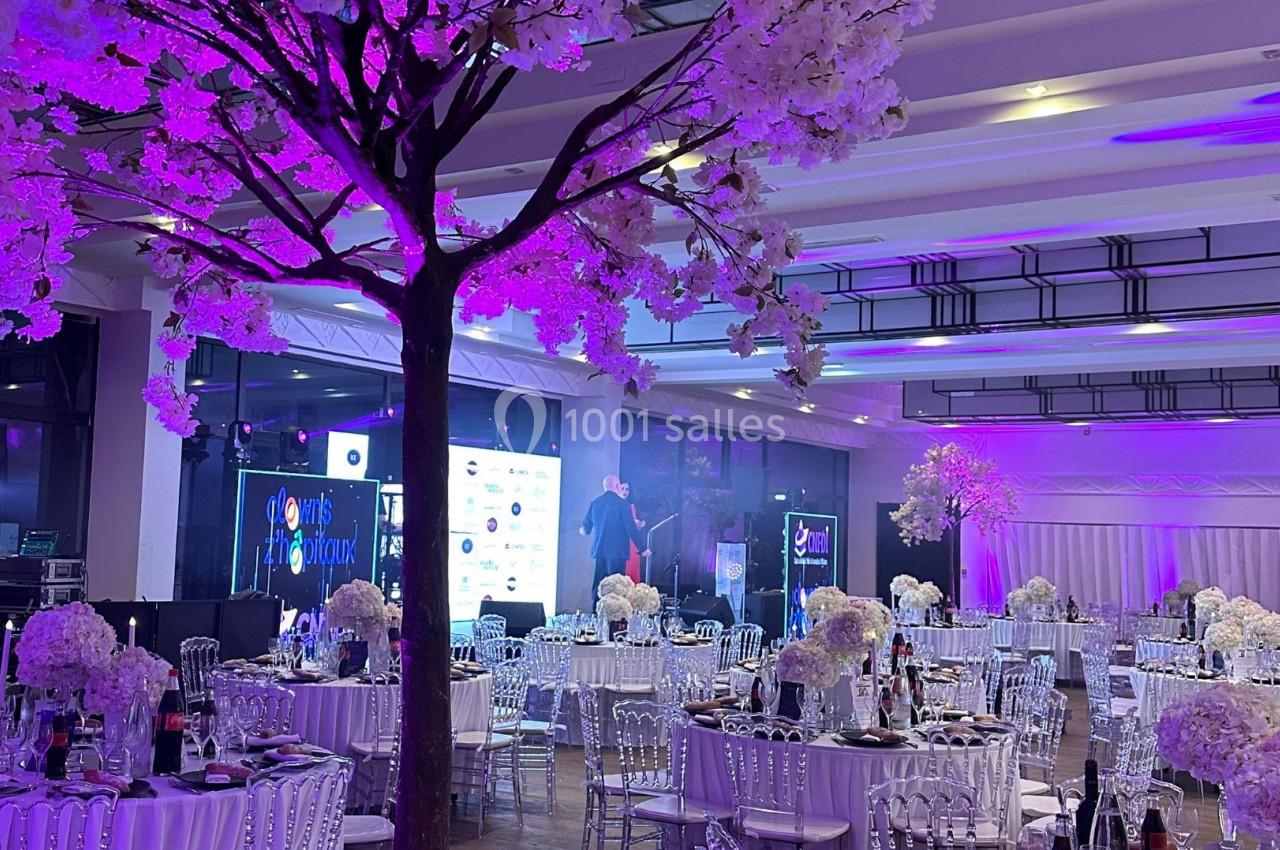 Salle de réception décorée avec des tables élégantes, des fleurs blanches et un éclairage violet.