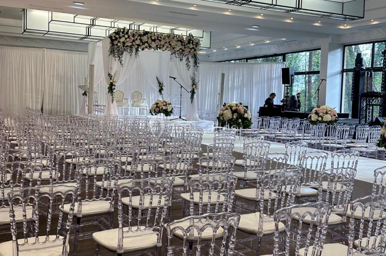 Salle de réception décorée pour une cérémonie, avec des chaises transparentes alignées et une arche florale blanche.