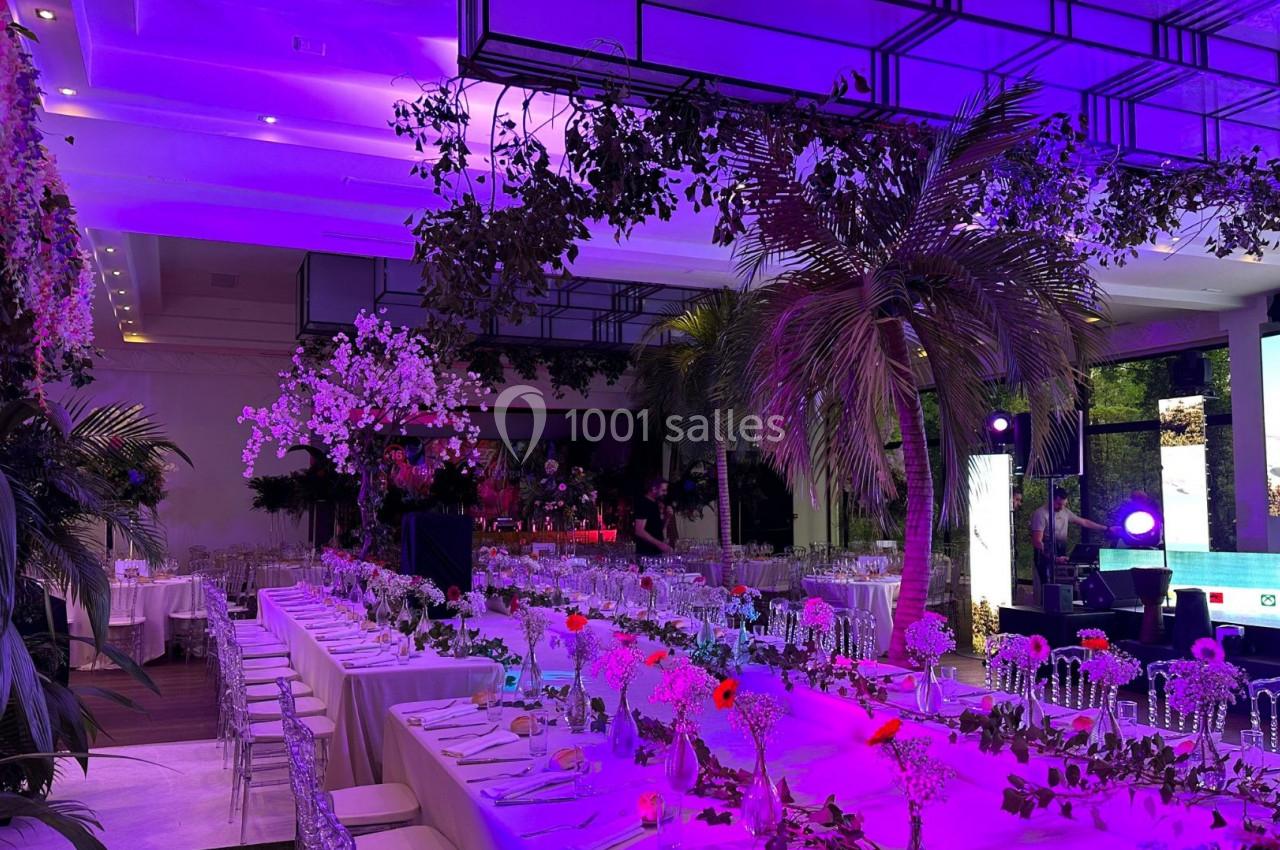 Salle de réception décorée avec des tables élégantes, des fleurs, des palmiers et un éclairage violet.