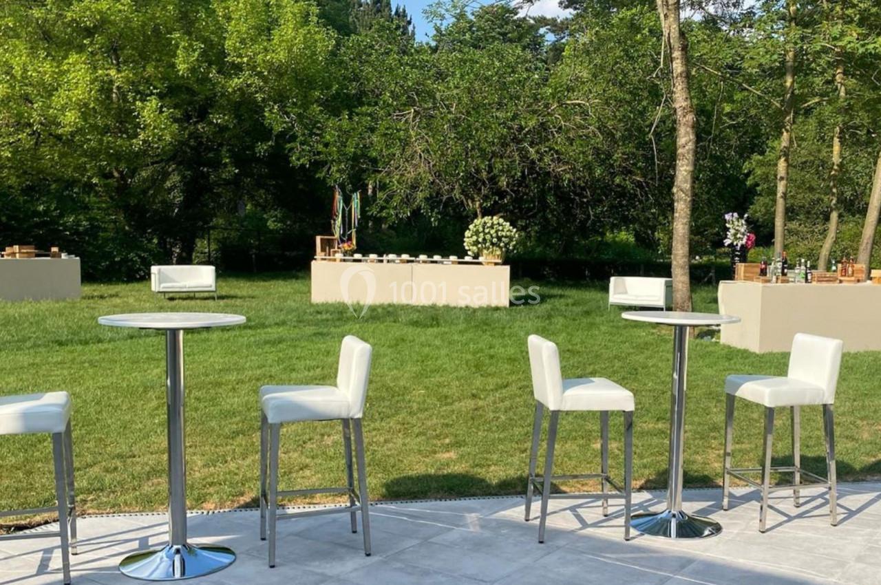 Tables hautes et chaises blanches disposées sur une terrasse, avec des buffets en arrière-plan dans un jardin verdoyant.