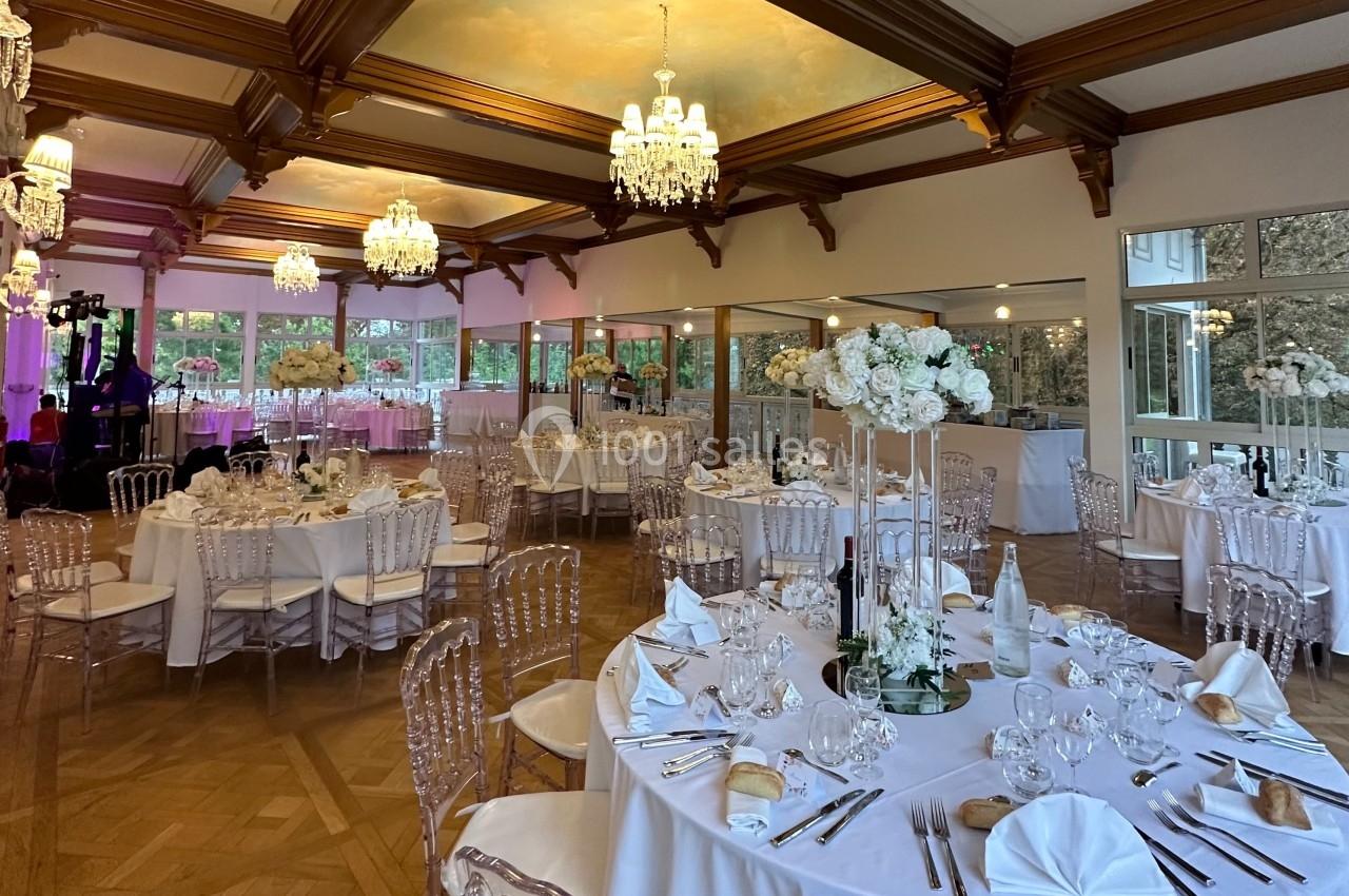 Salle de réception élégante avec tables dressées, chaises transparentes, décor floral blanc et lustres suspendus.