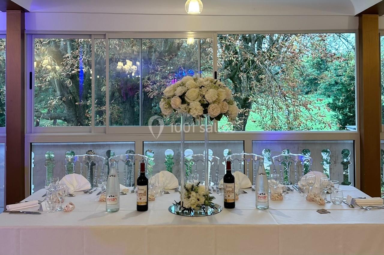 Table élégamment dressée avec des fleurs blanches, des bouteilles de vin et une vue sur un jardin à travers de grandes baies…