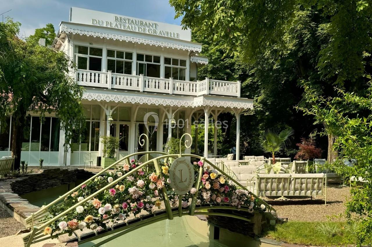 Façade d'un restaurant élégant entouré de verdure, avec un pont fleuri au premier plan.